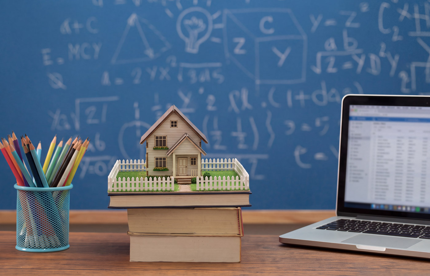 MCS-Phoenix-home-remodel-General-Contractor-sm2 Miniature house on stacked books sits beside colorful pencils and an open laptop, against a blue chalkboard of math formulas, suggesting learning.